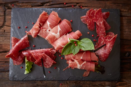 Slices of Italian prosciutto crudo or jamon with fresh basil leaves on a black background. Mediterranean appetizer or antipasto. Raw ham. Top view. Copy space.