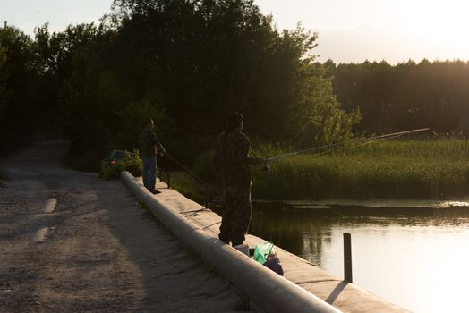 Silhouettes of two people fishing on a fishing rod with bright sunlight early in the morning at sunrise.