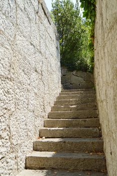 Old narrow staircase with stone wall on both sides, which leads up, illuminated by sunlight.