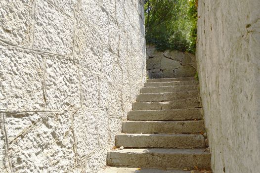 Old narrow staircase with stone wall on both sides, which leads up, illuminated by sunlight.