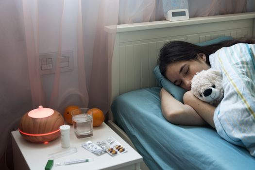 The young girl has a cold, lies in bed at home, feels unwell, takes medicines and vitamins, sleeps with a toy bear.