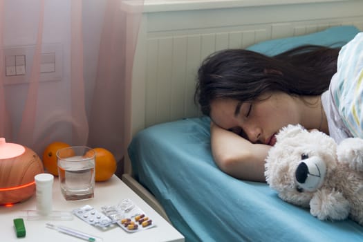 The young girl has a cold, lies in bed at home, feels unwell, takes medicines and vitamins, sleeps with a toy bear.