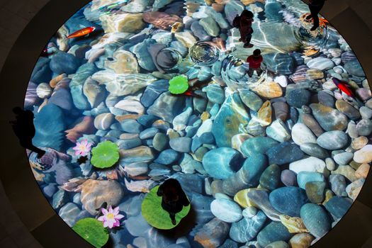 The large screen of augmented reality is felt on the floor. Digital Light Canvas, people with their children have fun on screen with a picture of the river bottom with beautiful stones and fish that swim away when you step on them.