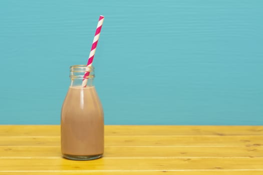 Chocolate milkshake with a retro paper straw in a one-third pint glass milk bottle, on a wooden table against a teal background