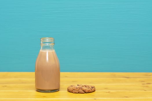 Chocolate milkshake in a retro one-third pint glass milk bottle and a chocolate chip cookie, on a wooden table against a bright teal painted background