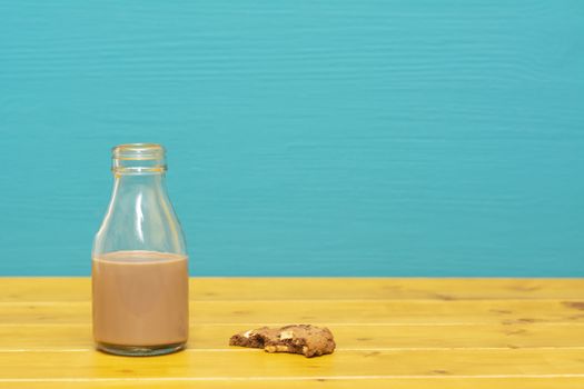 One-third pint glass milk bottle half full with chocolate milkshake and a half-eaten chocolate chip cookie, on a wooden table against a teal background