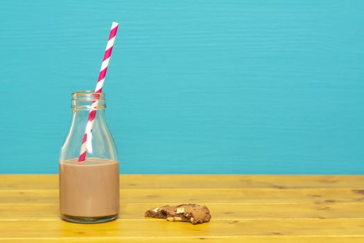 One-third pint glass milk bottle half full with chocolate milkshake with a retro straw and a half-eaten chocolate chip cookie, on a wooden table against a teal background