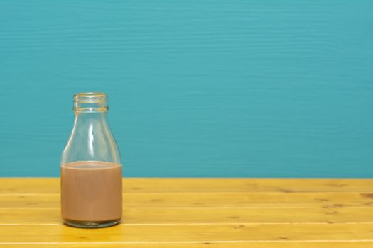 One-third pint glass milk bottle half full with chocolate milkshake, on a wooden table against a bright teal painted background