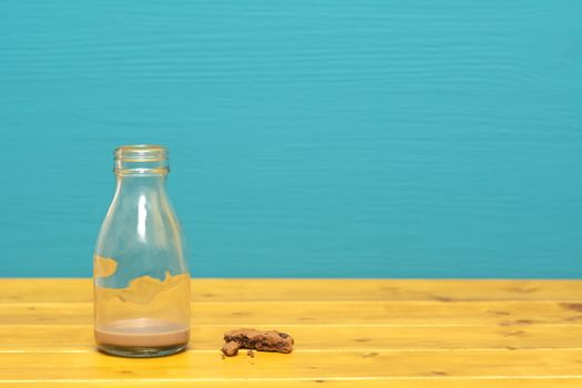 One-third pint glass milk bottle with dregs of chocolate milkshake and chocolate chip cookie crumbs, on a wooden table against a teal background