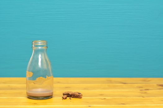 One-third pint glass milk bottle with dregs of chocolate milkshake and chocolate chip cookie crumbs, on a wooden table against a teal background