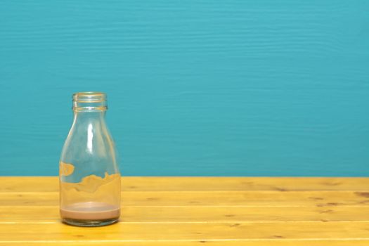 One-third pint glass milk bottle with dregs of chocolate milkshake, on a wooden table against a bright teal painted background