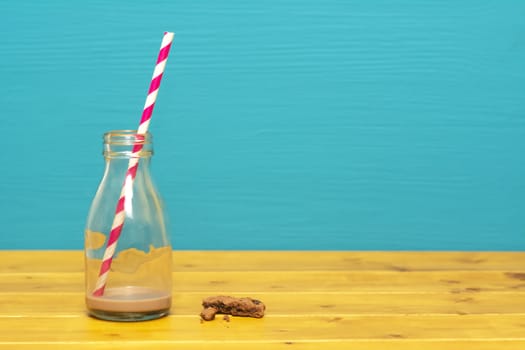 One-third pint glass milk bottle with dregs of chocolate milkshake with a retro straw and a chocolate chip cookie crumbs, on a wooden table against a teal background