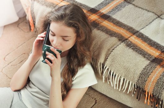 Cheerful brunette girl talking on mobile phone while sitting on comfortable couch. Smiling hipster girl having cell telephone conversation while resting on sofa in home interior and drinks coffee