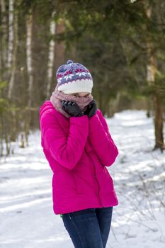 Portrait of a teenage girl in a bright pink winter jacket and knitted hat, which covers her face with a scarf. She stay in the winter snowy forest.