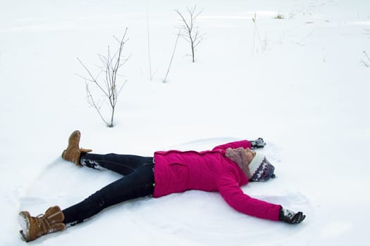 Portrait of a teenage girl in a bright pink winter jacket and knitted hat in the winter in the open air, she lies on pure white snow and makes a snow angel