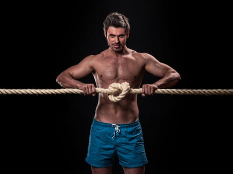 Attractive muscular man holding node of heavy ropes on black background