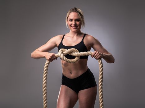 Attractive muscular woman holding node of heavy ropes on gray background