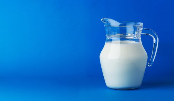 Glass jug of fresh milk isolated on blue background with copy space, pitcher of cream