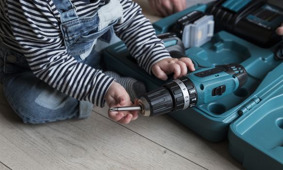 a one  year old child sitting on the ground putting a new drill in a drilling machine  which is located in a blue plastic case