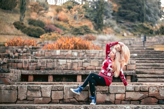 A woman in casual clothes is resting sitting on stone steps in an autumn park and straightens her long blond hair.