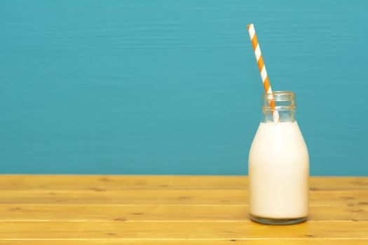Fresh creamy milk with a retro paper straw in a one-third pint glass milk bottle, on a wooden table against a teal background