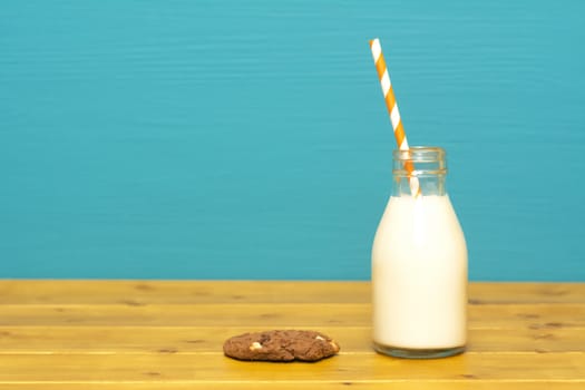 Fresh creamy milk with a retro paper straw in a one-third pint glass milk bottle and a chocolate chip cookie, on a wooden table against a teal background