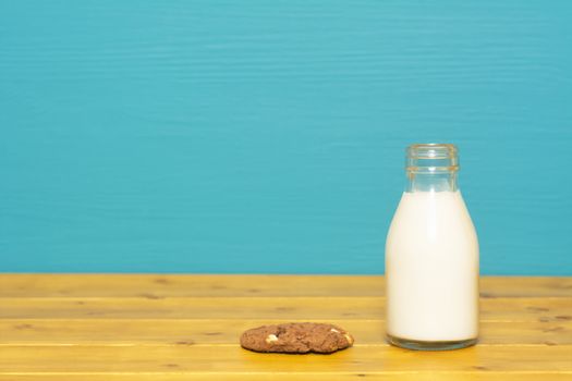 Fresh creamy milk in a one-third pint glass milk bottle and a chocolate chip cookie, on a wooden table against a bright teal painted background
