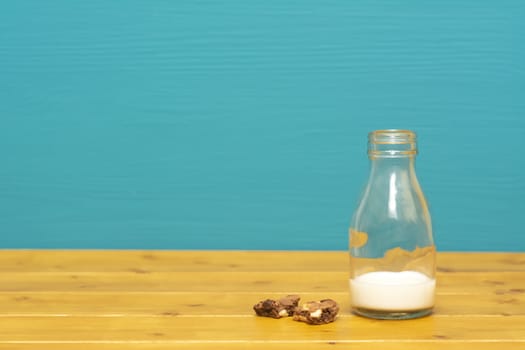 One-third pint glass milk bottle half full with fresh creamy milk and chocolate chip cookie crumbs, on a wooden table against a teal background