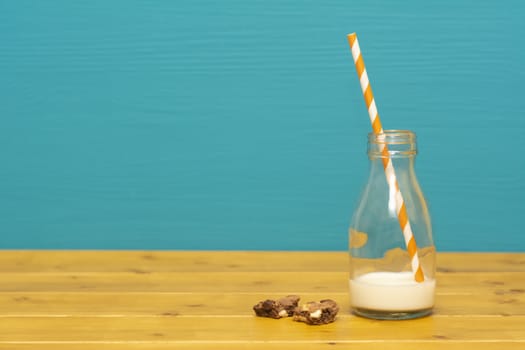 One-third pint glass milk bottle half full with fresh creamy milk with a retro straw and a chocolate chip cookie crumbs, on a wooden table against a teal background