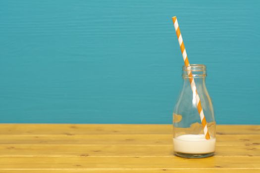 One-third pint glass milk bottle with dregs of fresh creamy milk with a retro paper straw, on a wooden table against a teal background