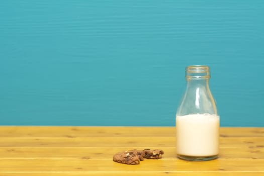 One-third pint glass milk bottle half full with fresh creamy milk and a half-eaten chocolate chip cookie, on a wooden table against a teal background