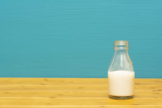 One-third pint glass milk bottle half full with fresh creamy milk, on a wooden table against a bright teal painted background