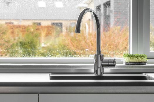 Chrome faucet by a kitchen sink with a wet window in the background