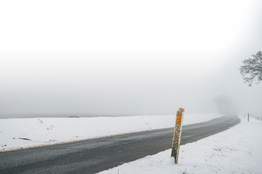 Marker post by a misty road in the winter with snow and a ice highway