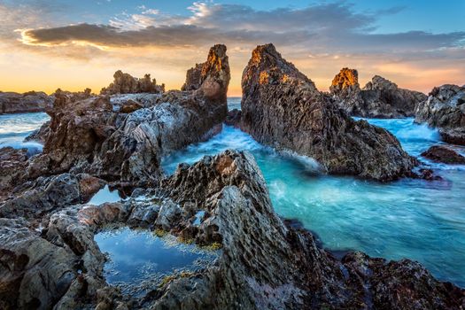 Unique rocky craggy pillars form a gully and channel the tidal waves through their narrow opening which widens out. In the foreground a rockpool edged with seaweed. The sun has just risen and its first warm rays are hitting the tips of the rocks
