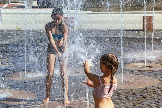 Children on a sunny summer day are poured water from a fountain.Children happily in shallow clean water on of city fountain on warm bright summer day.