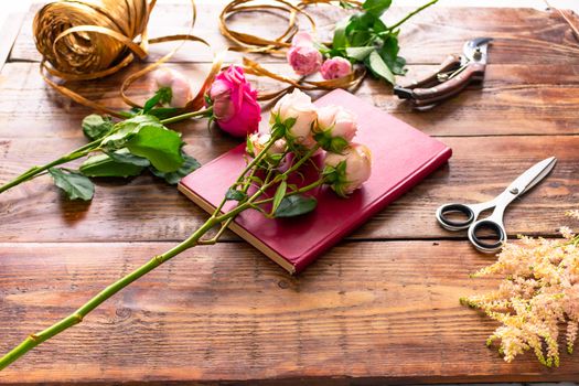 On the wooden surface laid out tools for flower bouquets: scissors, roses, colored ribbon, roses.