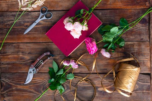 On the wooden surface laid out tools for flower bouquets: scissors, roses, colored ribbon, roses.