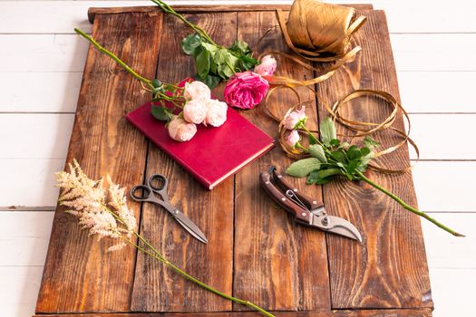 On the wooden surface laid out tools for flower bouquets: scissors, roses, colored ribbon, roses.