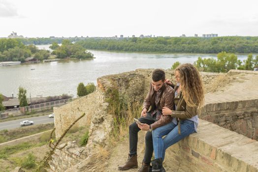 Attractive long haired woman and her boyfriend sitting on the wall and using their smart phone