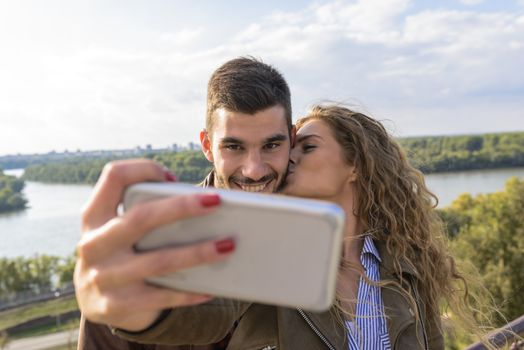 Beautiful young woman taking selfie in the nature while kissing her handsome boyfriend