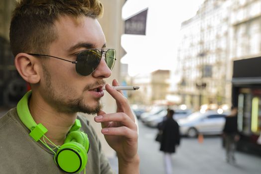 Young handsome man with green headphones and sunglasses enjoying a cigarette in the street.