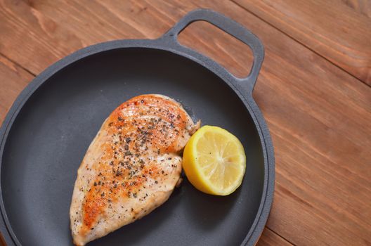 Fried chicken fillet is lying on a frying pan with a lemon on a wooden table