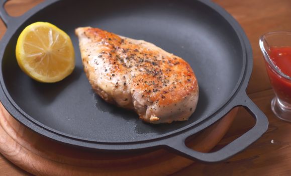 Large chicken fillet with lemon baked in a frying pan, next to the table there is a saucer