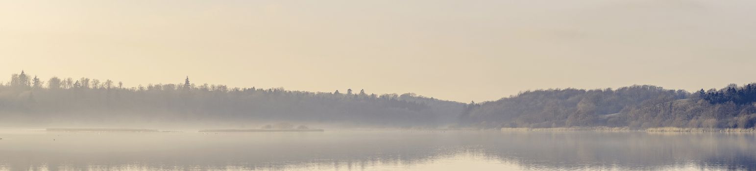 Panorama view over a misty lake in the morning sun with idyllic landscape scenery