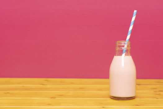 Strawberry milkshake with a retro paper straw in a one-third pint glass milk bottle, on a wooden table against a pink background
