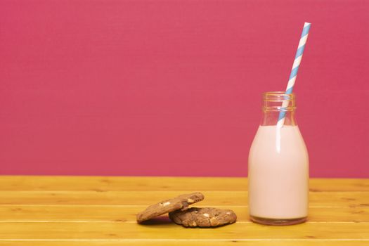 Strawberry milkshake with a retro paper straw in a one-third pint glass milk bottle and a chocolate chip cookie, on a wooden table against a pink background