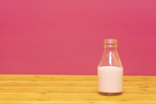 One-third pint glass milk bottle half full with strawberry milkshake, on a wooden table against a pink painted background