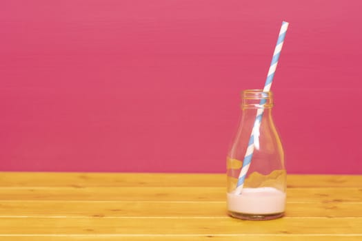 One-third pint glass milk bottle with dregs of strawberry milkshake and a retro paper straw, on a wooden table against a pink background