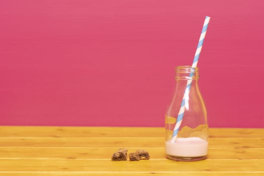 One-third pint glass milk bottle half full with strawberry milkshake with a retro straw and chocolate chip cookie crumbs, on a wooden table against a pink background